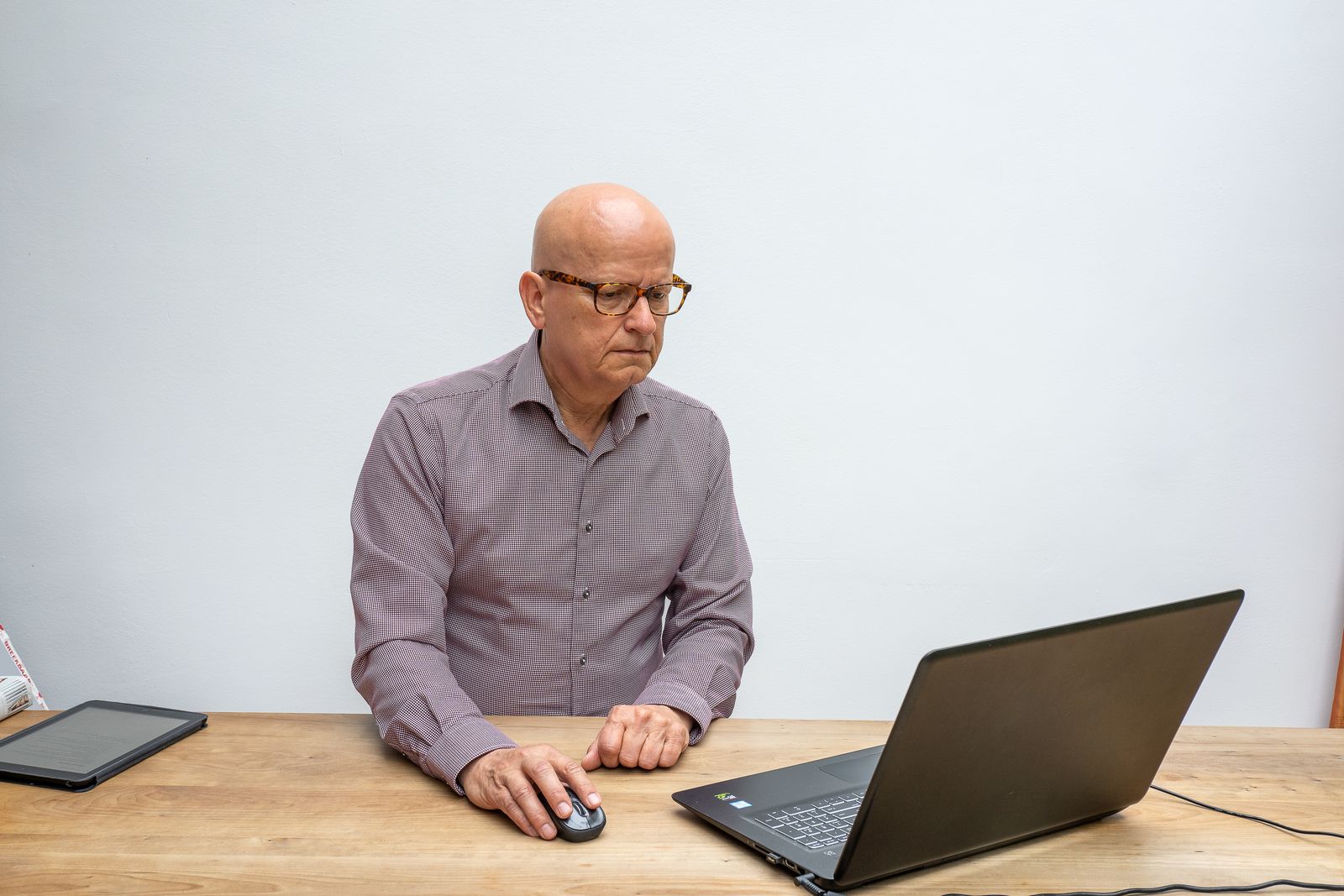 Elderly man using laptop and mouse, focused at work in a bright setting.