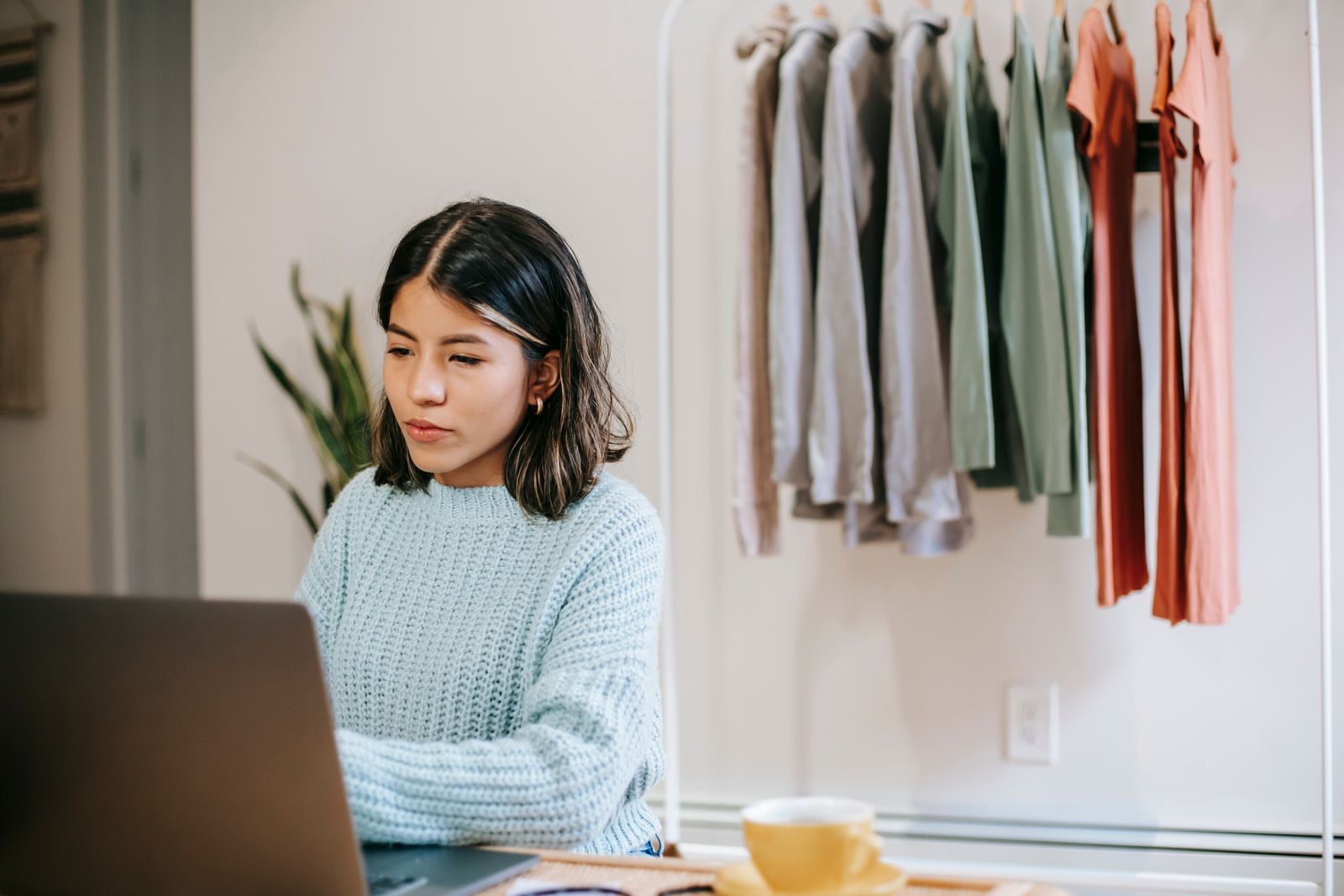 Serious young Hispanic female freelancer in casual outfit surfing on laptop at table near cup with drink at bright home with wardrobe with garments and plant with green leaves